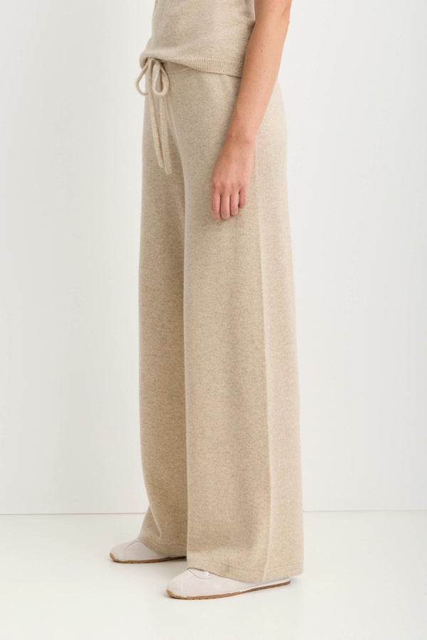 A person wearing loose, wide-leg knit pants in a light warm beige or oatmeal tone, paired with a matching top and pale slip-on shoes, photographed against a plain light background.